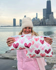 Woman carrying two zipper pouches with heart print with city skyline behind her.