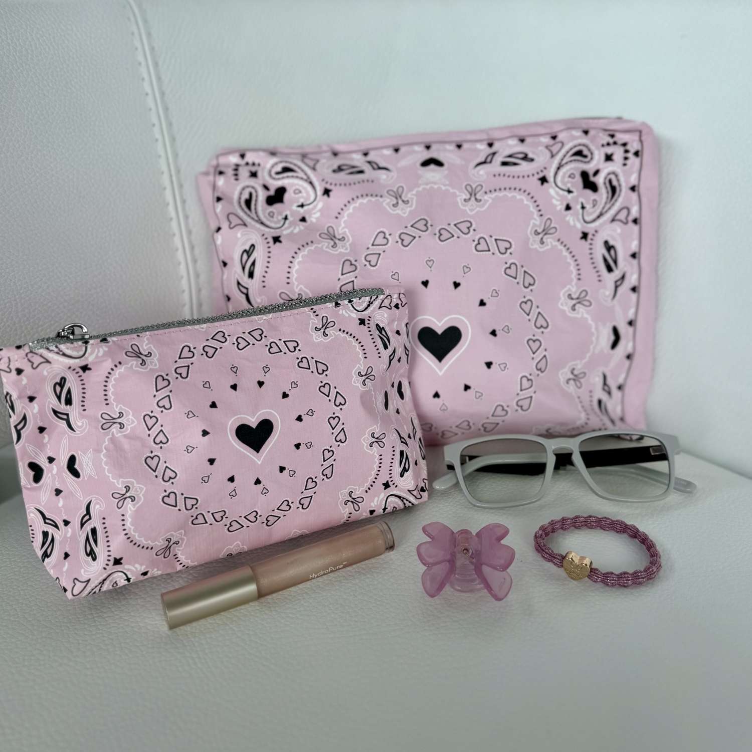 Pink pouches with black heart patterns, sunglasses, lip gloss, butterfly charm, and bracelet on a light surface.