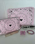 Pink pouches with black heart patterns, sunglasses, lip gloss, butterfly charm, and bracelet on a light surface.
