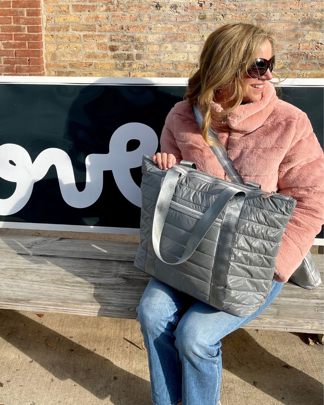 Woman sitting on bench in pink jacket, blue jeans, carrying puffer tote bag in cool gray
