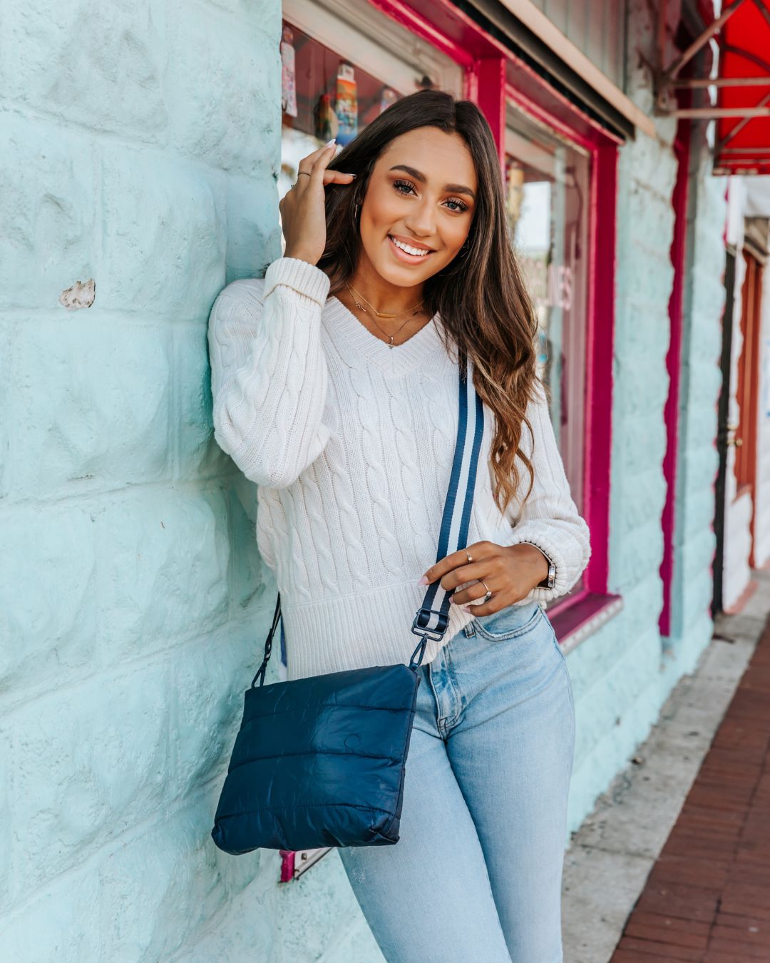 Woman in white sweater, jeans, and navy blue puffer crossbody purse standing against light blue wall