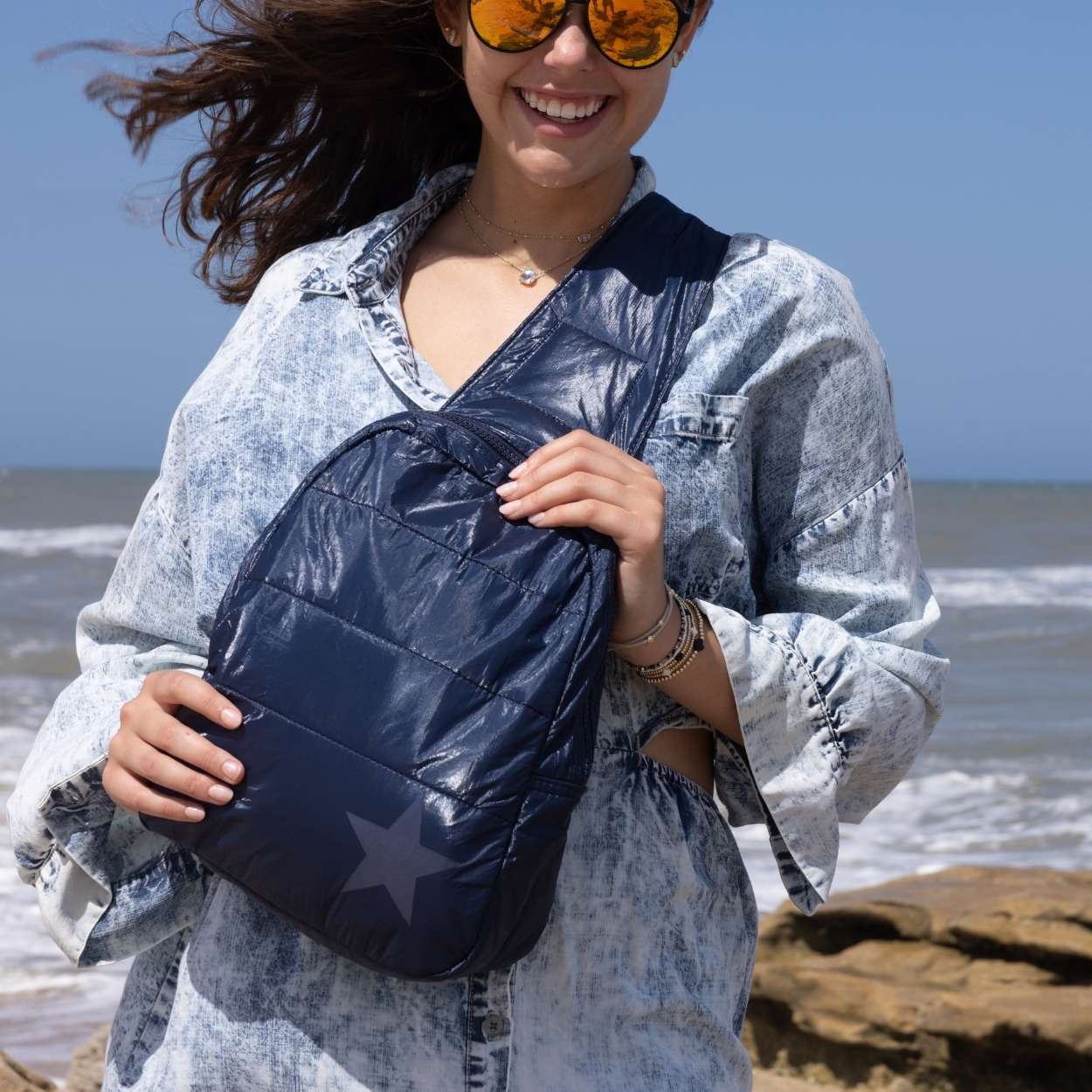 Woman at the beach holding crossbody backpack in shimmer navy with tonal star