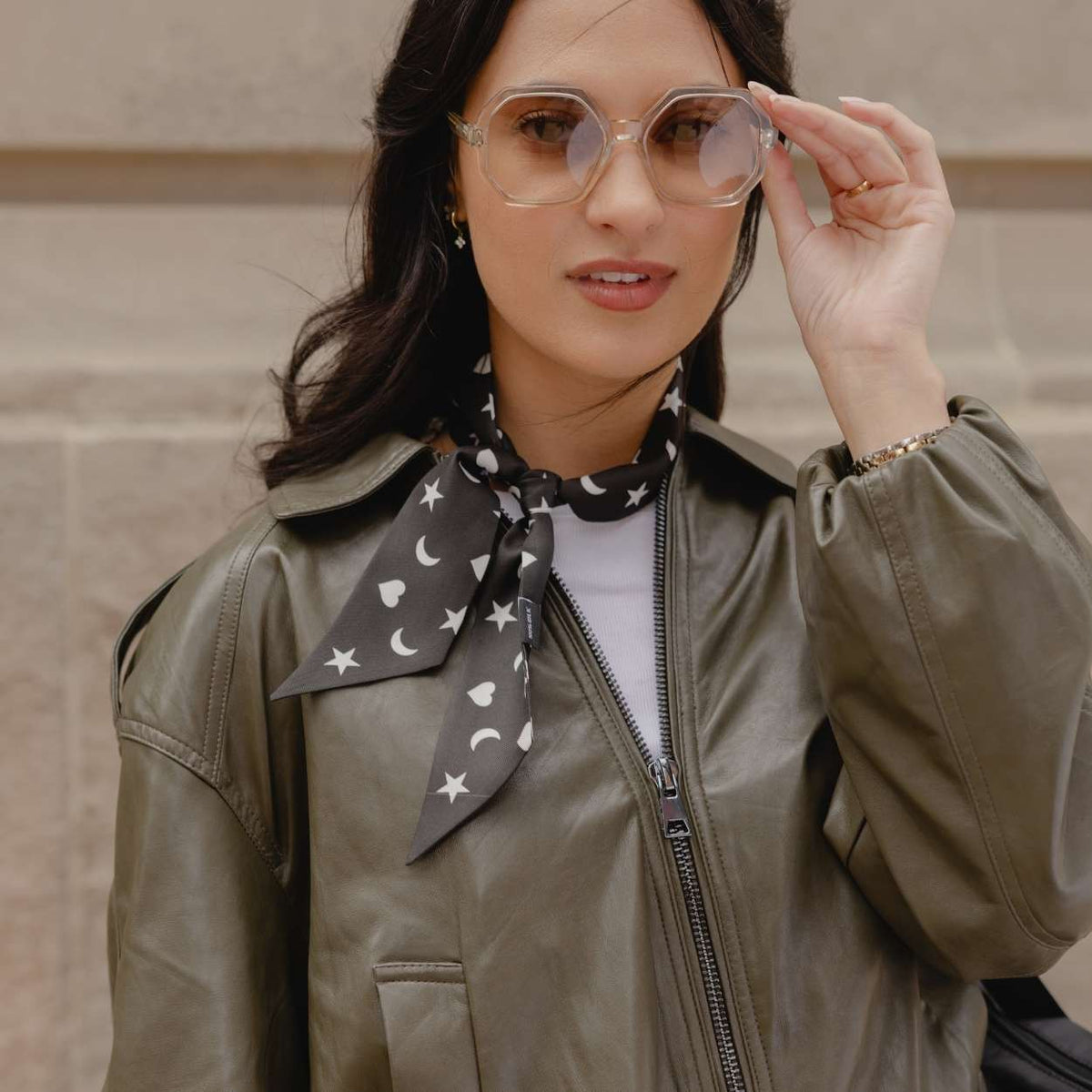 A woman wearing a black neck scarf with a white crescent moon, heart and star pattern, styled with an olive green leather jacket and clear-framed glasses.