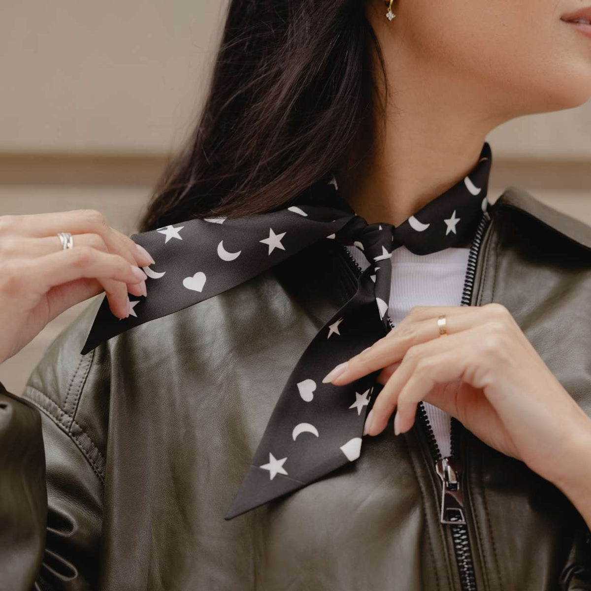 A close-up of a woman wearing a black satin scarf with a white celestial pattern of stars, hearts and crescent moons, styled under a dark olive green leather jacket.