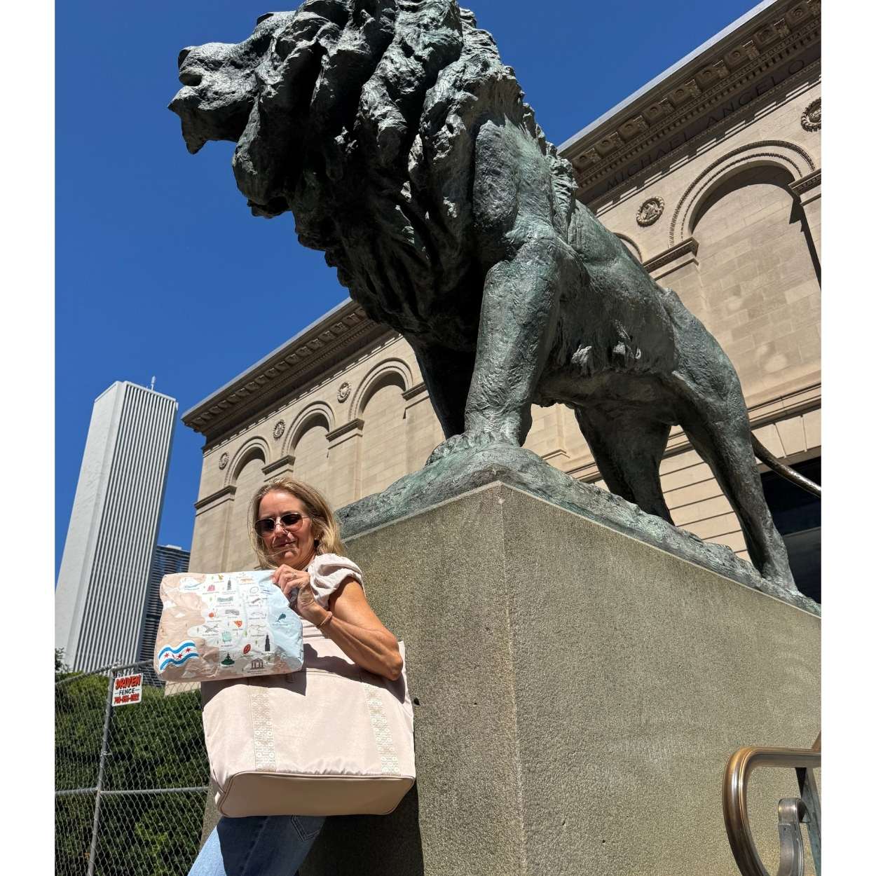 A woman stands outdoors beside a large bronze lion statue on a stone pedestal, holding a large beige tote bag and pouch with colorful illustrated Chicago landmarks. A beige building and blue sky are visible in the background.