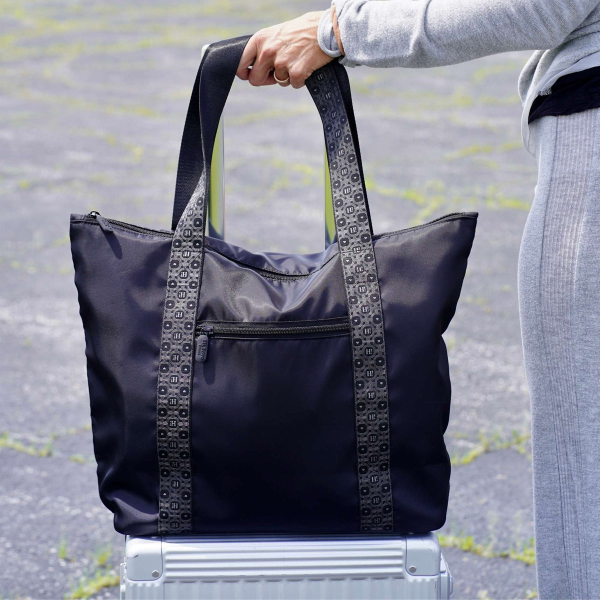 A black tote bag with signature patterned handles, held by a person standing on a silver suitcase outdoors on asphalt.