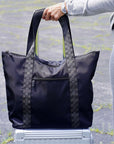A black tote bag with signature patterned handles, held by a person standing on a silver suitcase outdoors on asphalt.