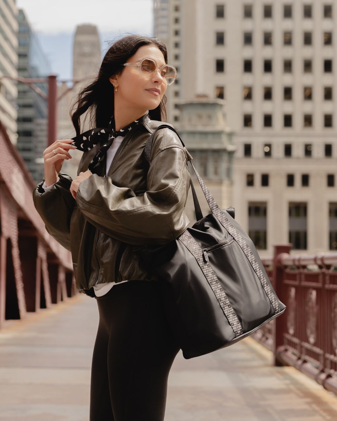 A woman wearing a shiny dark brown leather jacket, black leggings, and sunglasses carries a large black tote bag with a patterned strap over a Chicago city bridge.