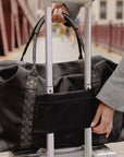 A black duffel bag with Hi Love Signature pattern on handles and trim, shown on a silver rolling suitcase with a person's hands holding the handles in an outdoor setting.