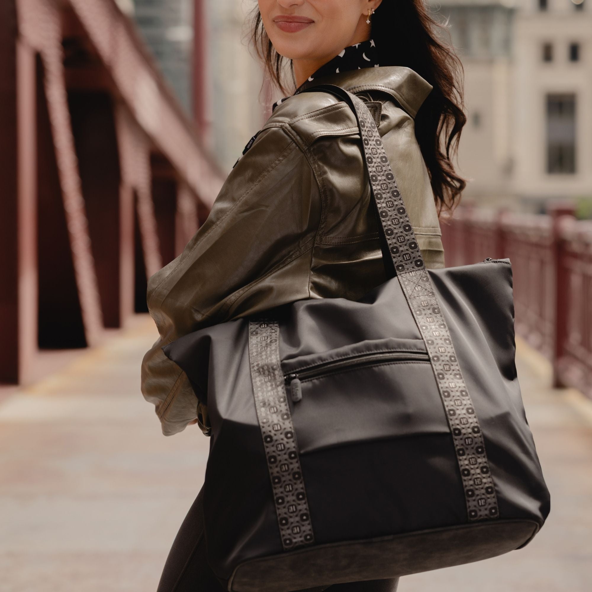 Woman in an olive jacket carrying the black signature tote over her shoulder on a city street.