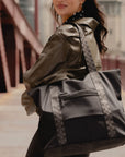 Woman in an olive jacket carrying the black signature tote over her shoulder on a city street.