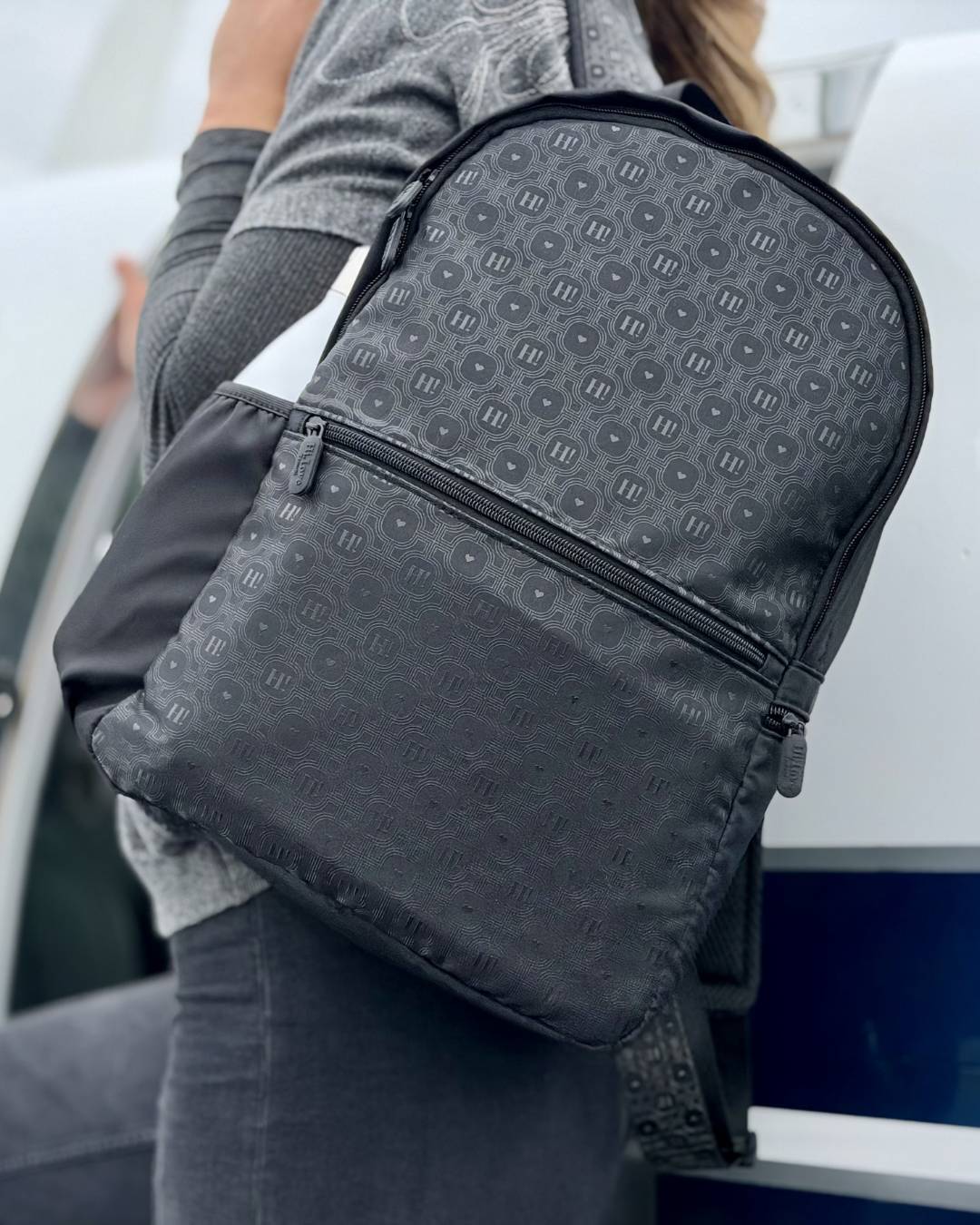 A black patterned backpack with a repeating monogram design, shown being worn by a person in a gray long-sleeve top and dark pants.