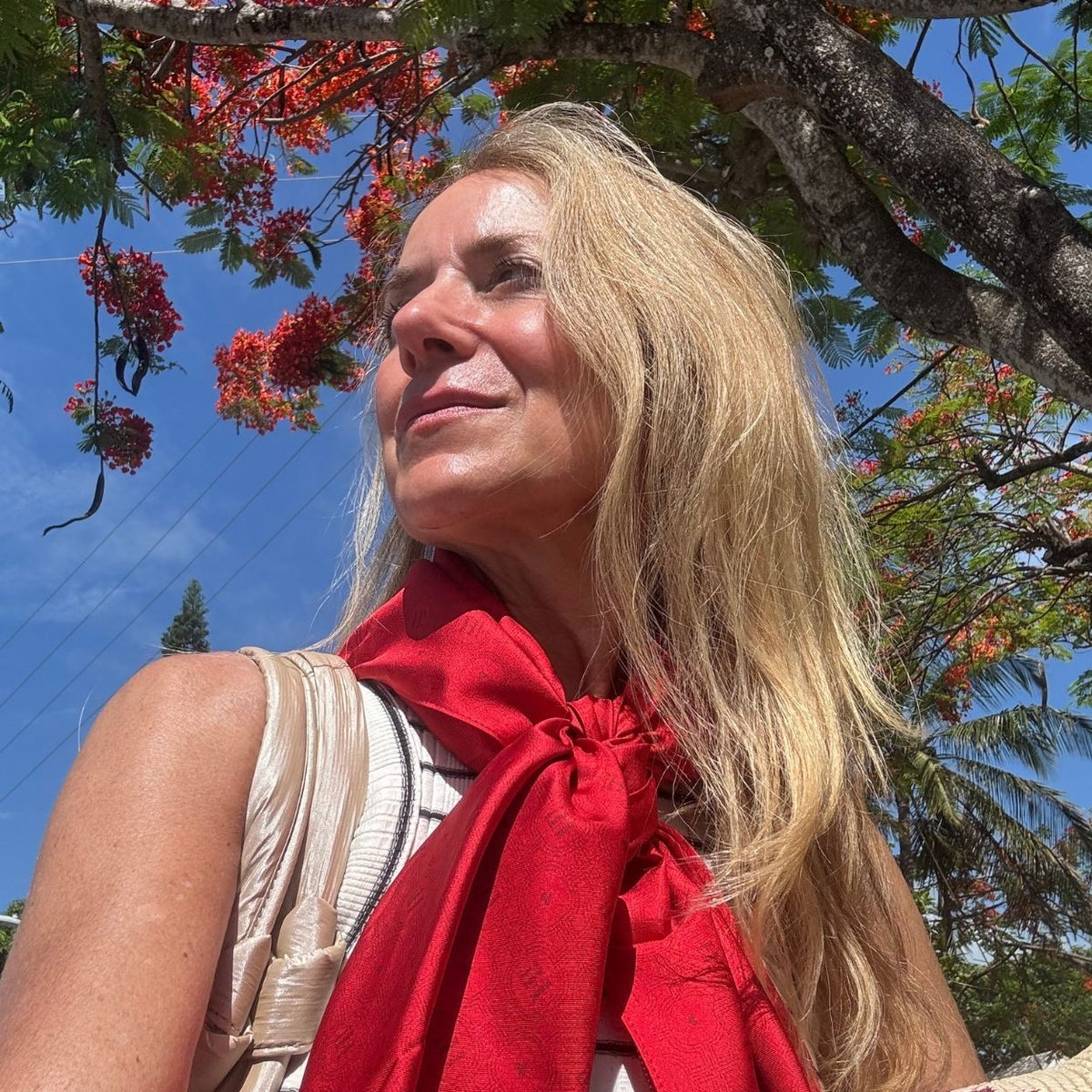 A woman outdoors under a tree wearing a red silk scarf with a subtle tonal "Hi" print on the scarf.