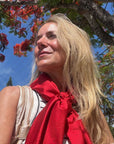 A woman outdoors under a tree wearing a red silk scarf with a subtle tonal "Hi" print on the scarf.