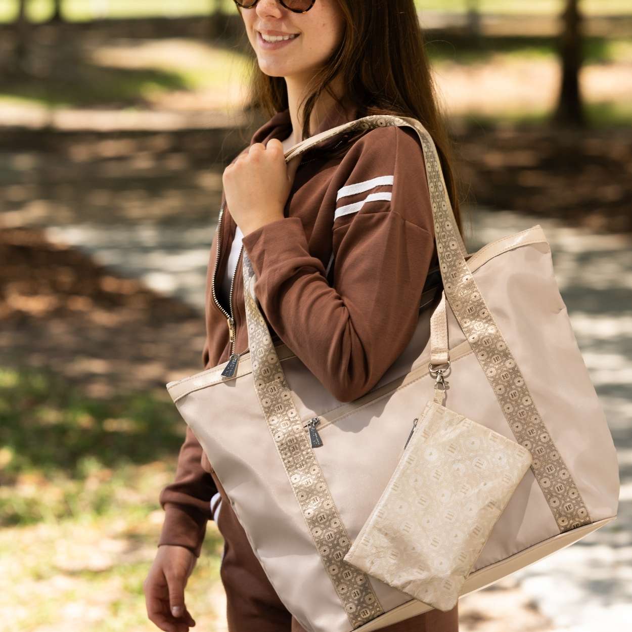 A woman outdoors carrying a large beige tote bag with patterned trim and a matching zip pouch, wearing a brown tracksuit.