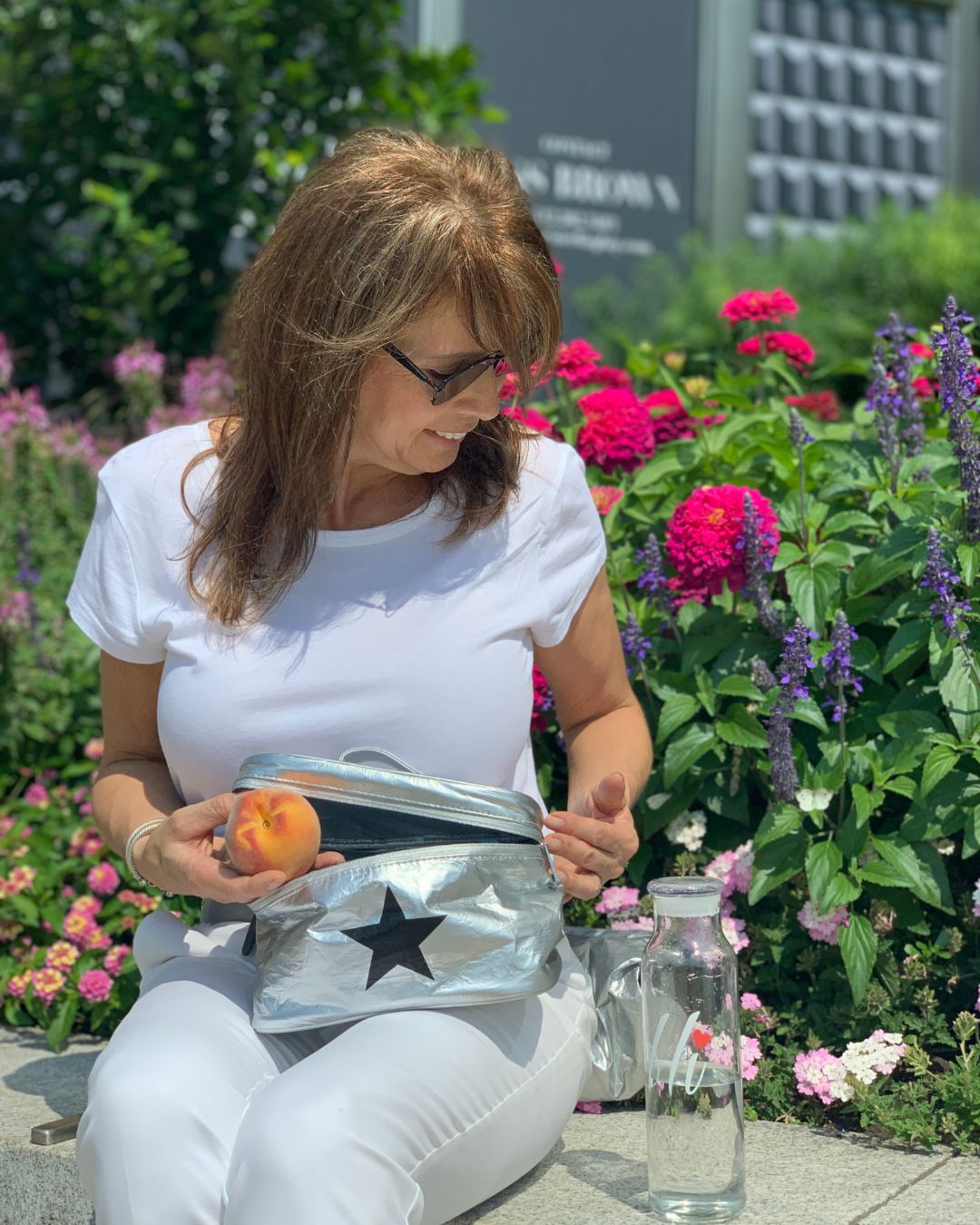 Woman in white outfit sitting holding silver lunch box with black star in outdoor floral setting