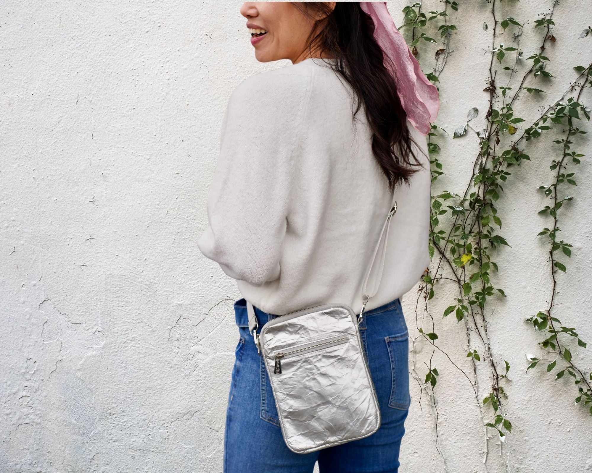 Woman wearing crossbody purse in silver against a cream wall with greenery 