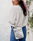 Woman wearing crossbody purse in silver against a cream wall with greenery 