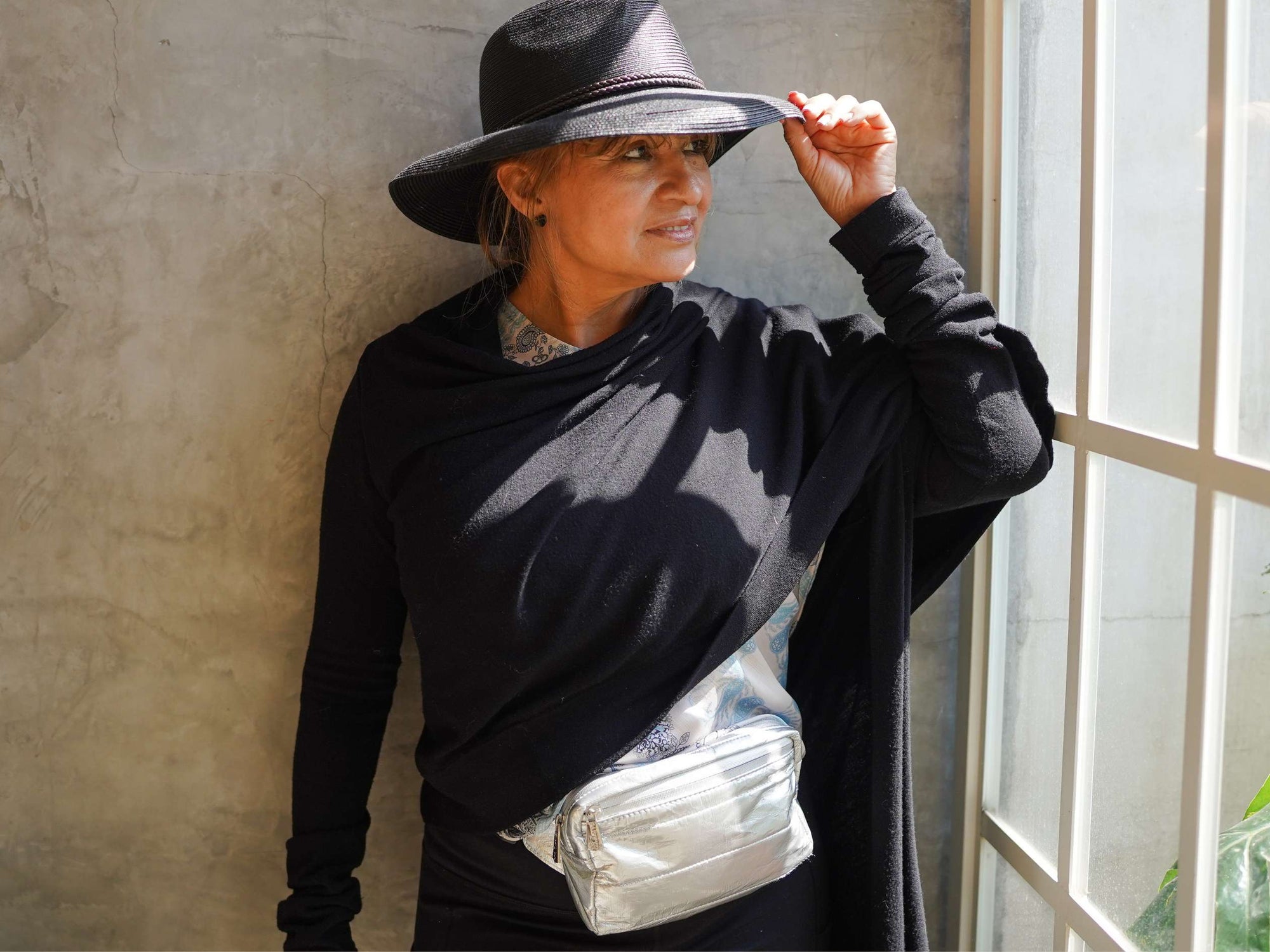 Person wearing a black outfit with a silver fanny pack, standing against a neutral wall.