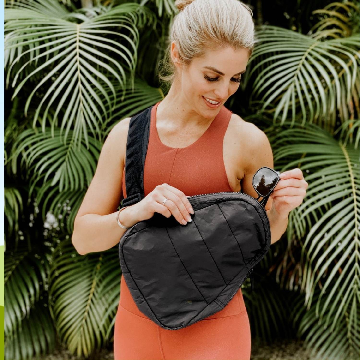 A woman wearing a rust-orange athletic tank top and matching shorts holds a black sling bag across her torso, with sunglasses in one hand and tropical palm leaves in the background.