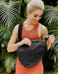 A woman wearing a rust-orange athletic tank top and matching shorts holds a black sling bag across her torso, with sunglasses in one hand and tropical palm leaves in the background.
