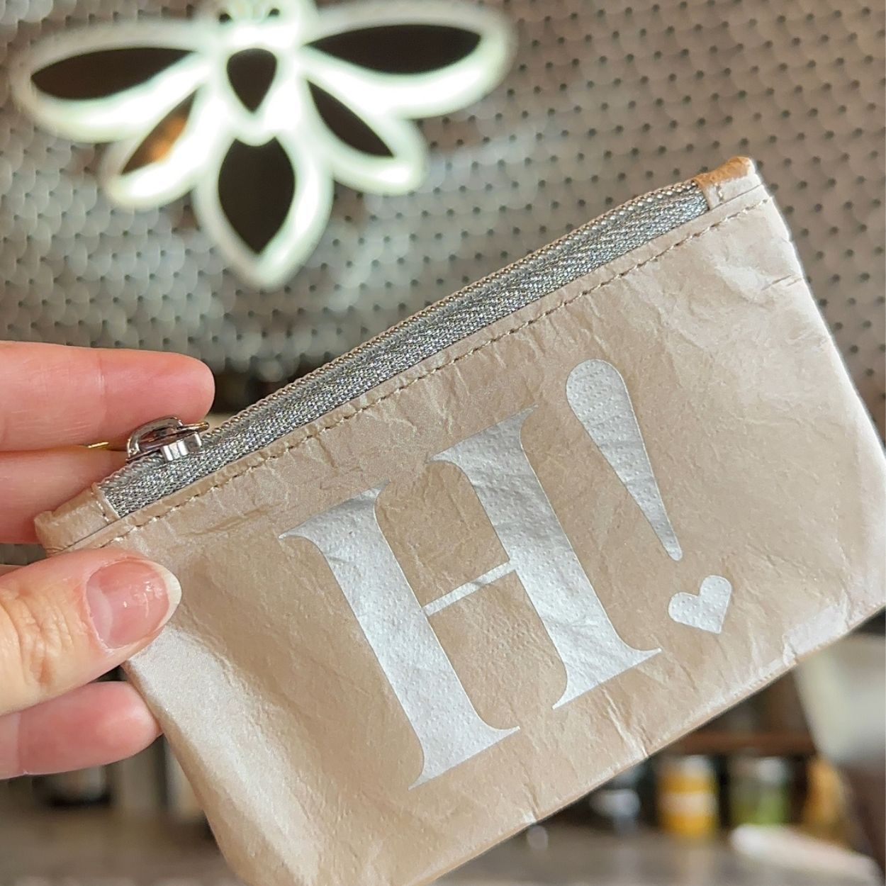 A close-up of a hand holding a small beige zip pouch with a silver zipper and a large white 'H!' printed on the front, set against a softly blurred background.