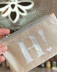 A close-up of a hand holding a small beige zip pouch with a silver zipper and a large white 'H!' printed on the front, set against a softly blurred background.