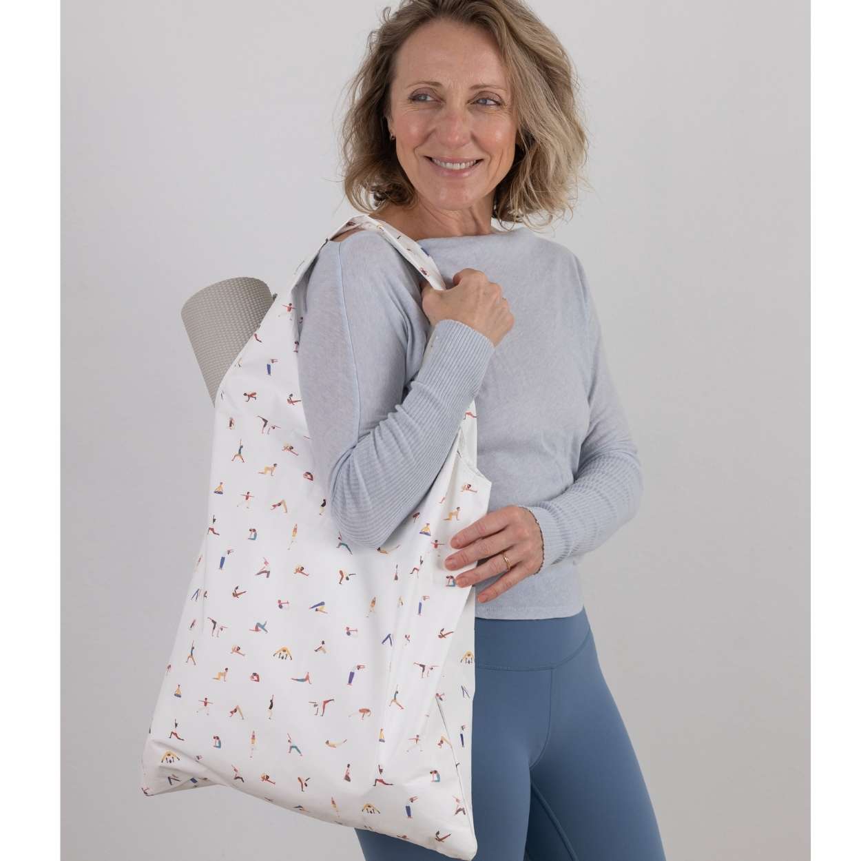 Woman holding a white tote bag with colorful yoga pose pattern against a plain background