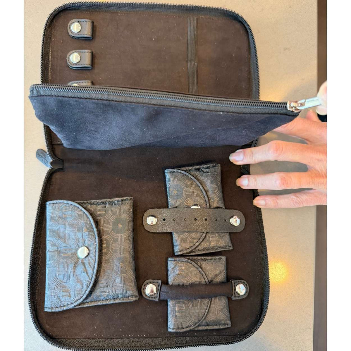 An open black zippered travel jewelry organizer case with multiple compartments, including a coin pocket, card holder, and strap details, shown with a hand holding the zipper tab.