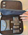 An open black zippered travel jewelry organizer case with multiple compartments, including a coin pocket, card holder, and strap details, shown with a hand holding the zipper tab.