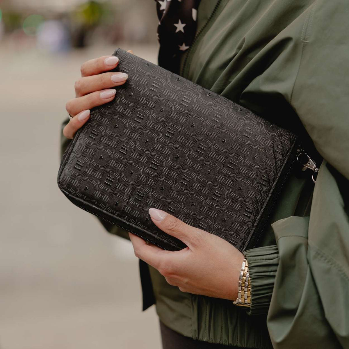 A close-up of a person holding a shimmer black textured clutch bag with a repeating embossed monogram pattern. The person is wearing an olive green jacket and a gold link bracelet, with a blurred outdoor background.
