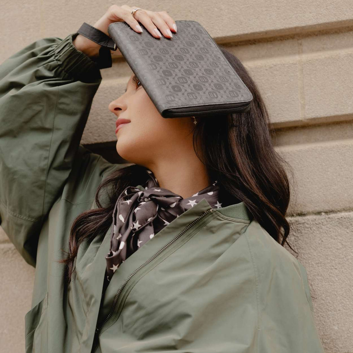 A woman in an olive green jacket and a patterned scarf holds a shimmer black monogram clutch bag over her eyes, posing against a beige stone wall.