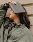 A woman in an olive green jacket and a patterned scarf holds a shimmer black monogram clutch bag over her eyes, posing against a beige stone wall.