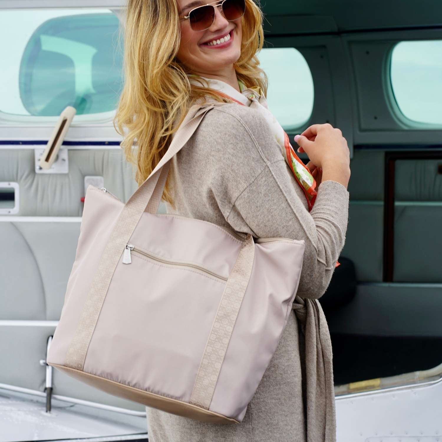 A woman wearing sunglasses and a beige cardigan carries a large, light beige zippered tote bag with a front zip pocket, set against the background of a private jet cabin.