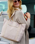 A woman wearing sunglasses and a beige cardigan carries a large, light beige zippered tote bag with a front zip pocket, set against the background of a private jet cabin.