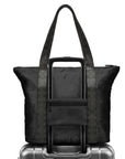 A black expandable tote with trolley sleeve with patterned straps, shown with its telescoping handle extended and resting on top of a silver suitcase against a white background.