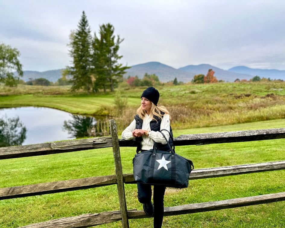 Woman standing in scenic field holding black duffle bag with silver star