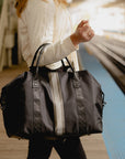 Black weekender bag with silver and pewter center stripes carried on the arm at a train platform.