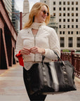 Woman carrying a black weekender bag with silver and pewter center stripes on a city bridge wearing a cream jacket and black pants.