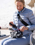 A woman sitting outdoors on a bench wearing a light blue puffer jacket and jeans, holding a small black zip wristlet pouch with white ski-print graphics. Another matching pouch rests on the table beside her.