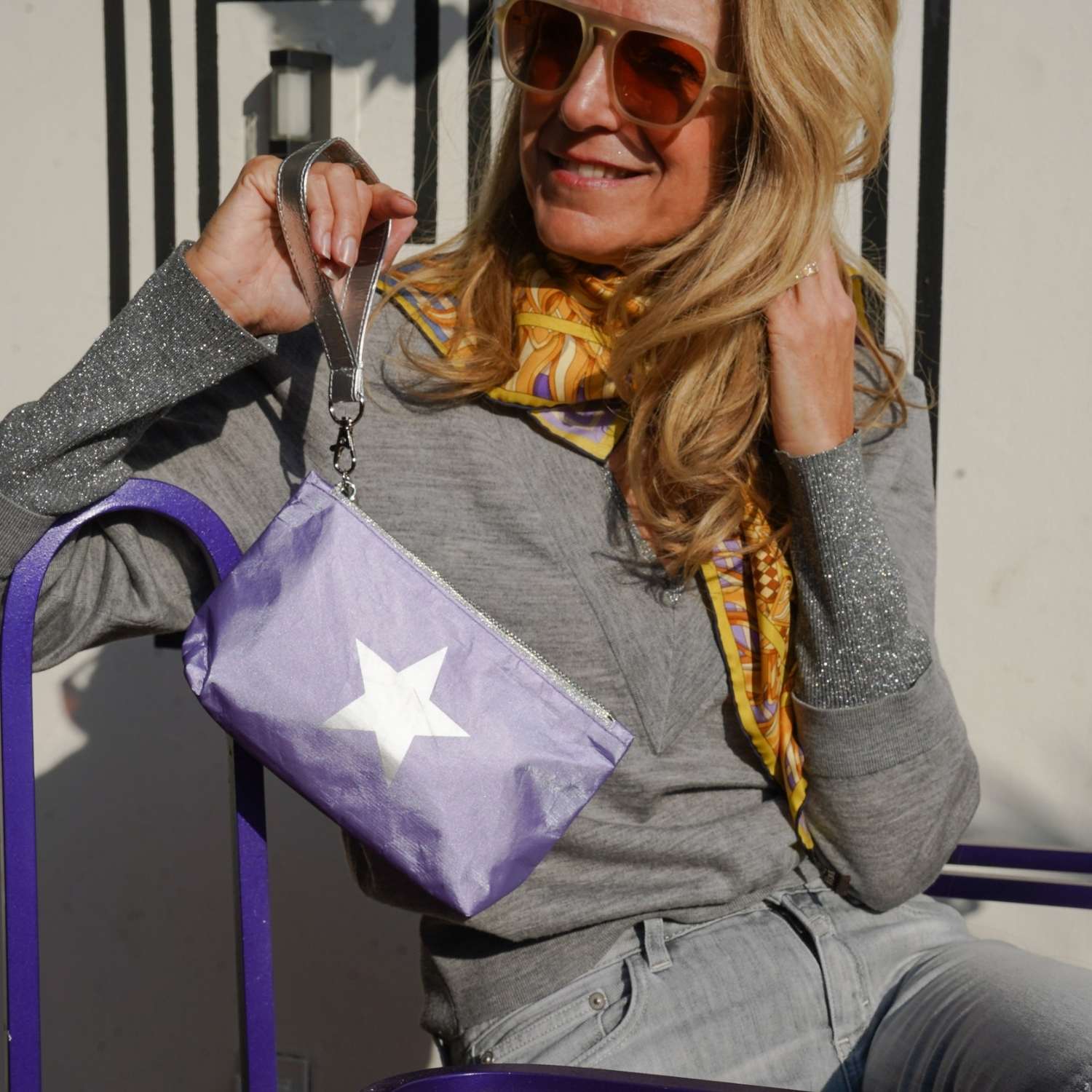 Woman holding a purple bag with a star design, wearing sunglasses and a patterned scarf.