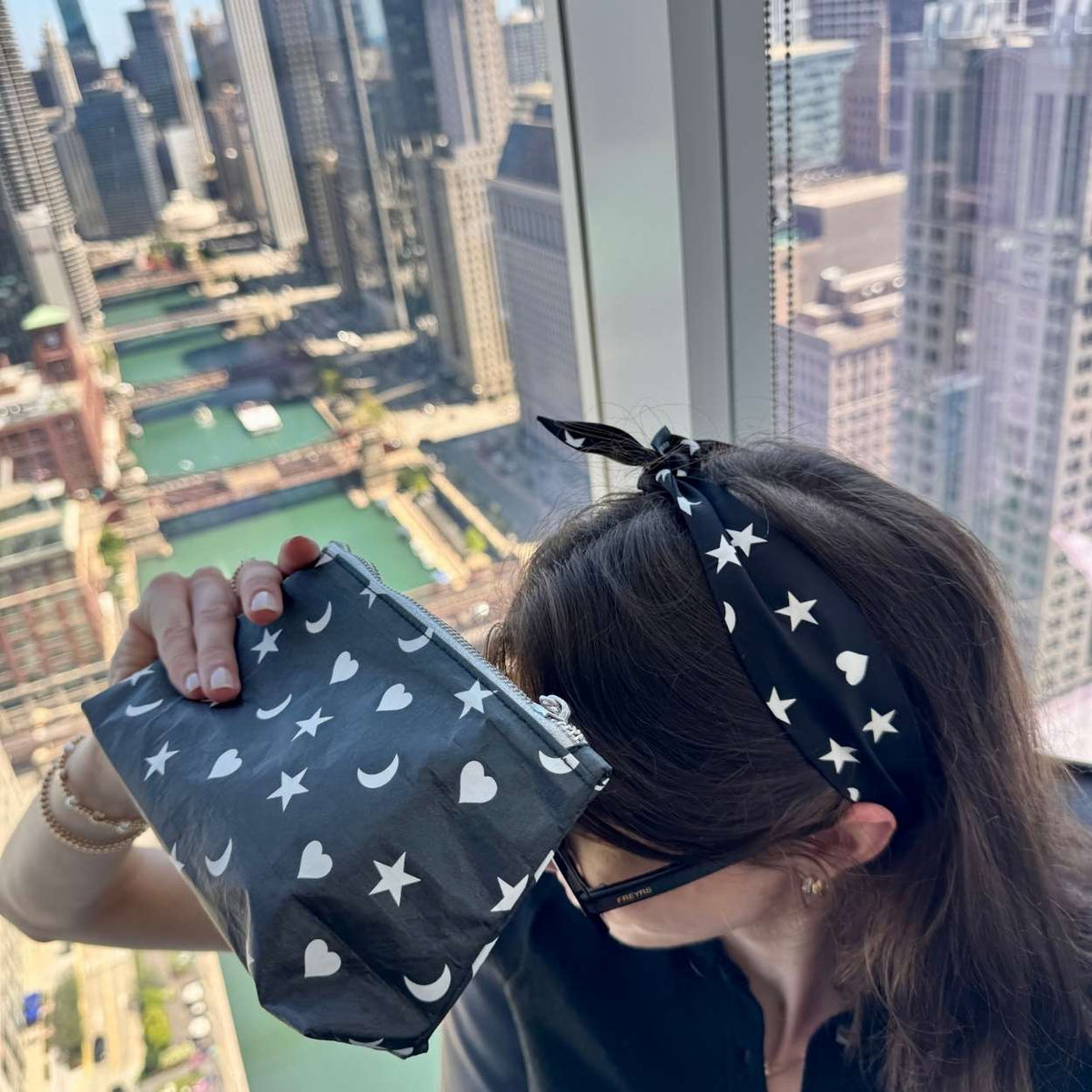 A person holding a black zippered pouch with a white star, heart and moon pattern, positioned in front of a large window with a city skyline view. The person is also wearing a matching black scarfette with white stars, hearts and moons.