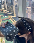 A person holding a black zippered pouch with a white star, heart and moon pattern, positioned in front of a large window with a city skyline view. The person is also wearing a matching black scarfette with white stars, hearts and moons.