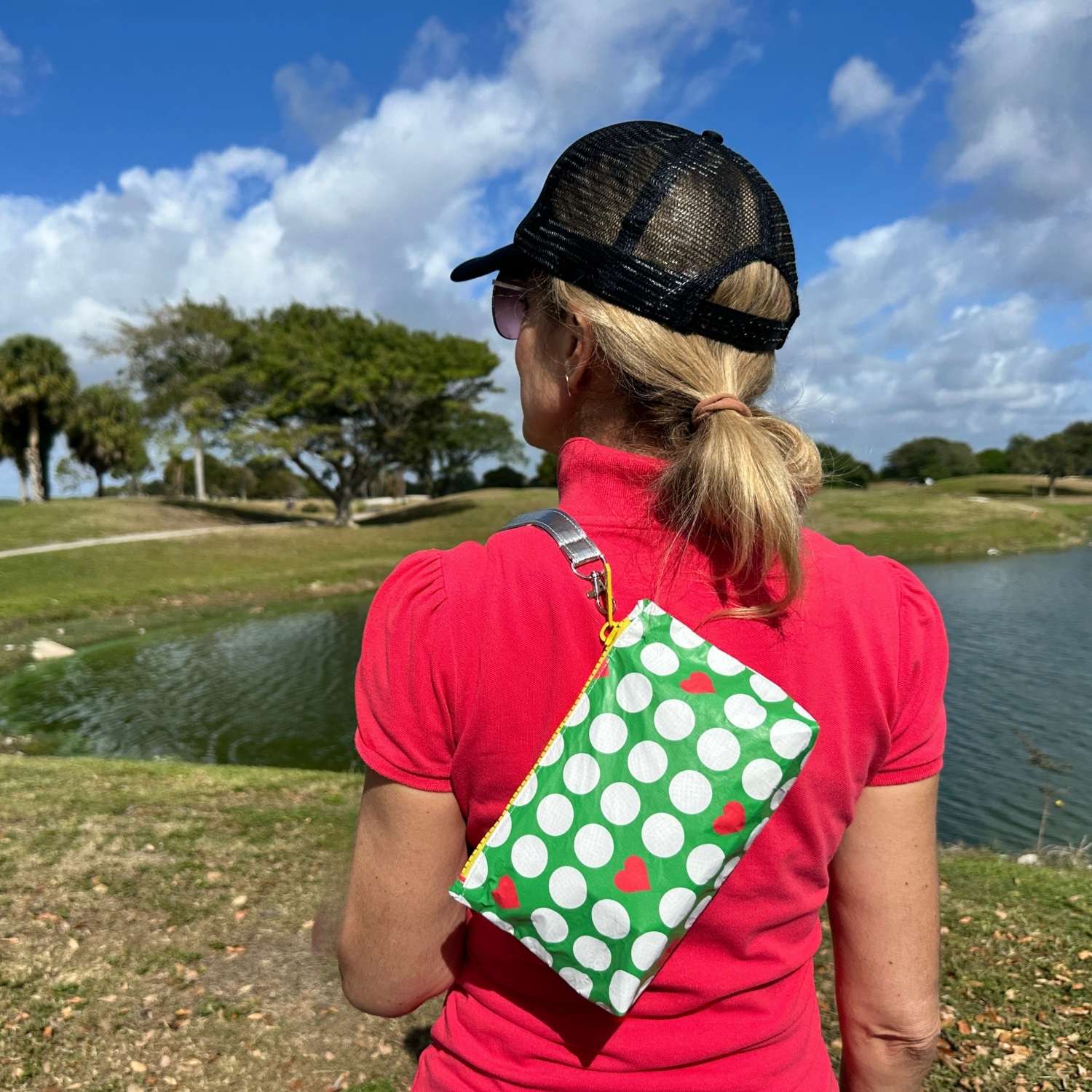 Person wearing a red shirt and black cap with a green wristlet with golf balls and red hearts, standing by a pond with trees in the background.