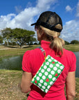 Person wearing a red shirt and black cap with a green wristlet with golf balls and red hearts, standing by a pond with trees in the background.