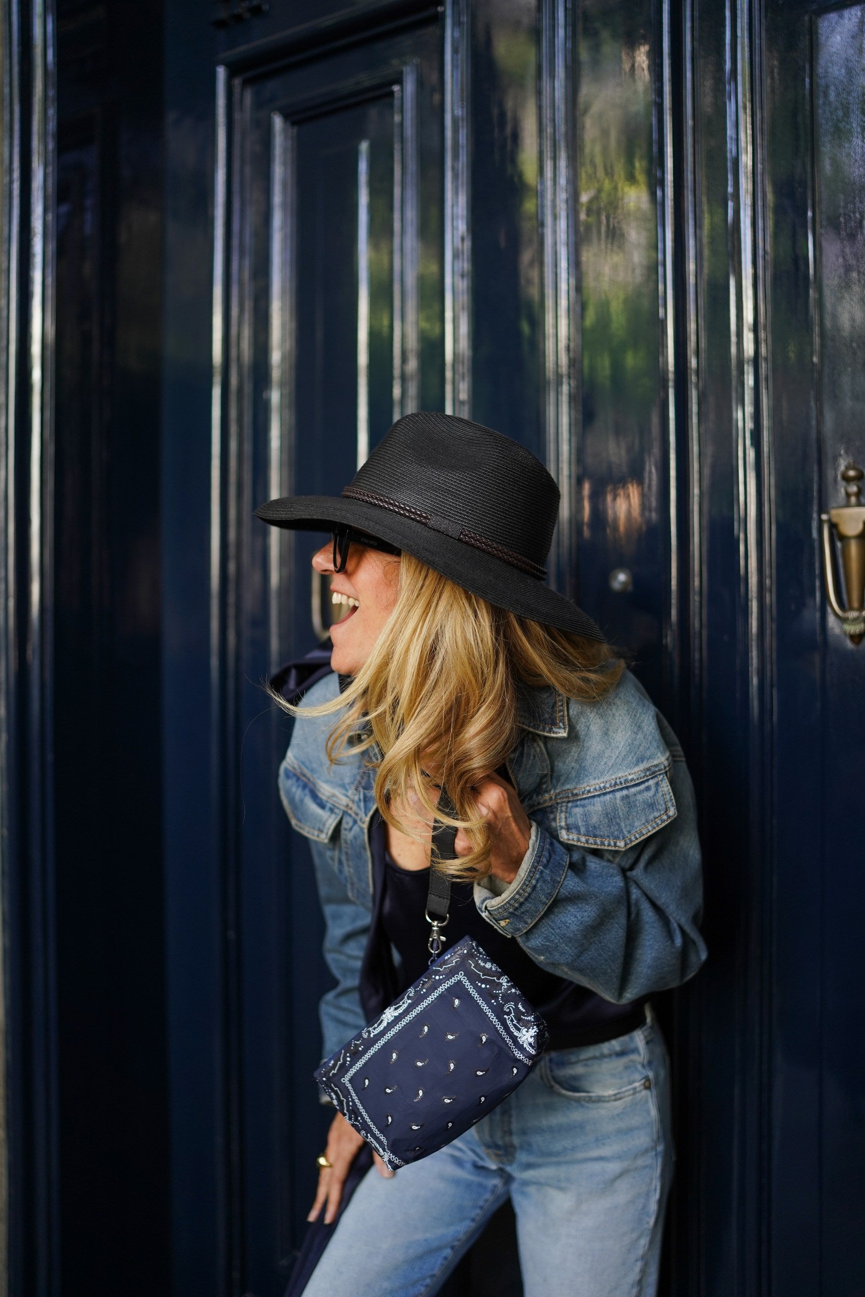 Woman wearing a navy blue bandana print wristlet with a black hat and denim jacket standing in front of a dark blue door.