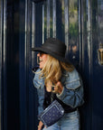 Woman wearing a navy blue bandana print wristlet with a black hat and denim jacket standing in front of a dark blue door.