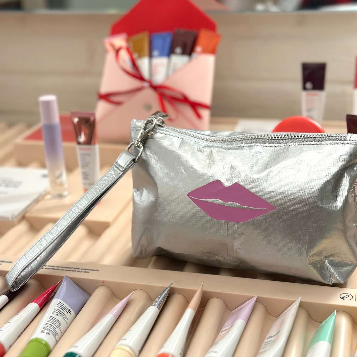 Silver makeup bag with pink lip design on a wooden surface with makeup items.