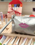 Silver makeup bag with pink lip design on a wooden surface with makeup items.