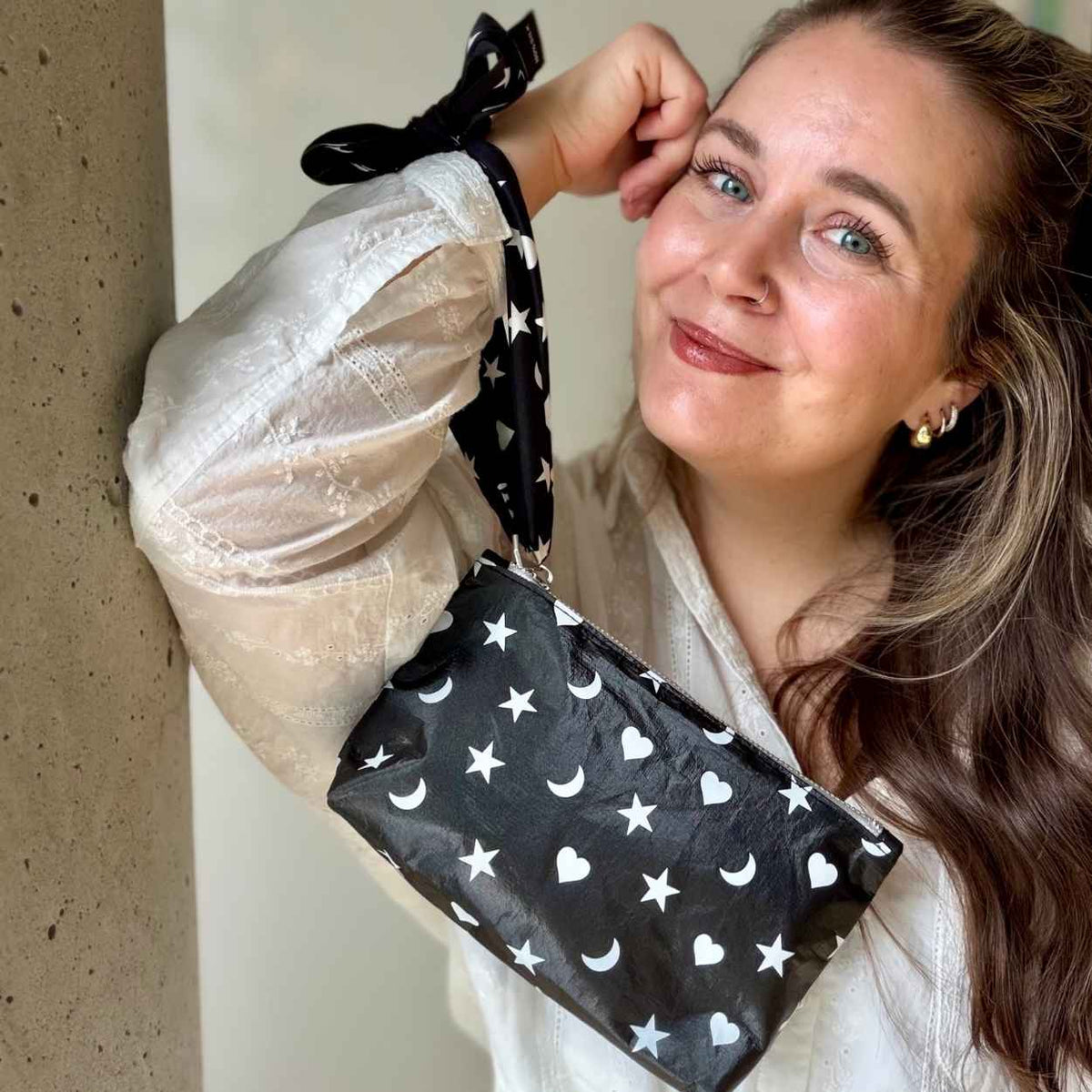 Woman wearing a black silk scarfette as a wristlet strap attached to a mini zipper pouch with white hearts, moons and stars pattern.